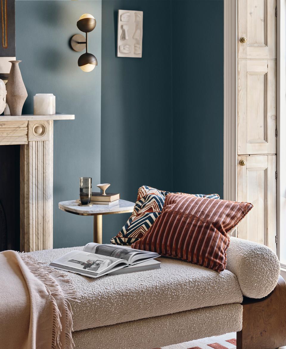 A chaise lounge with pillows and a blanket sits beside an open book and table with a glass, within a cozy room featuring a fireplace, wall art, and an open window with greenery outside.