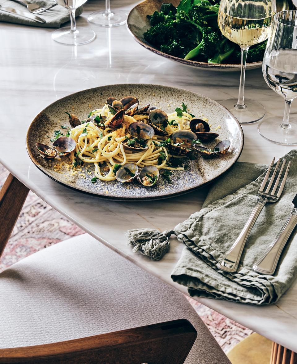 A ceramic plate with pasta and clams sits on a marble dining table with a cloth napkin, silverware, wine glasses, and a bowl of green vegetables in the background.