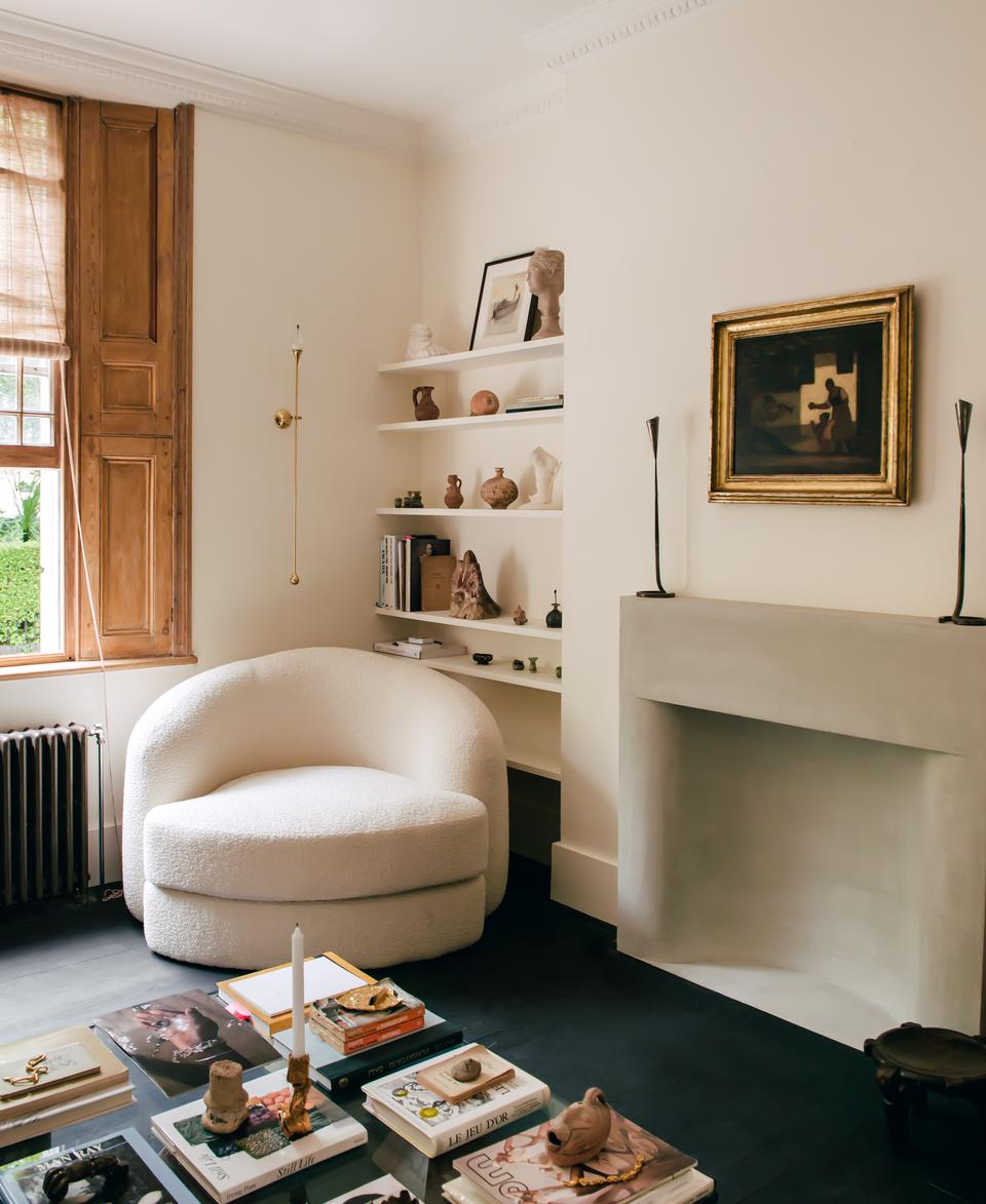 A white curved chair sits beside built-in shelves holding various art pieces and books in a cozy, beige-walled living room with wooden window shutters and a well-decorated fireplace.