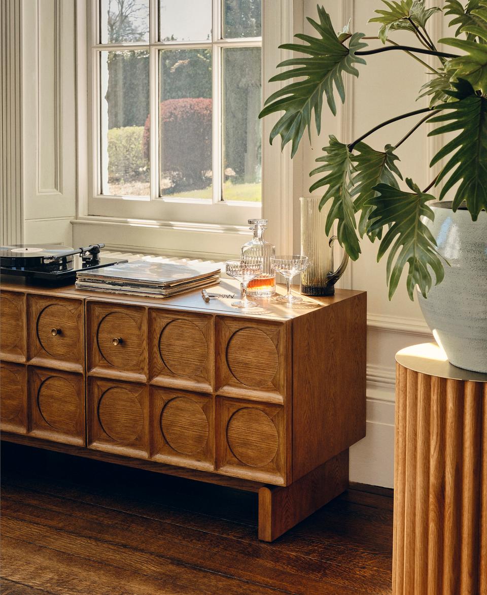 A wooden sideboard with geometric circular patterns holds vinyl records, a turntable, and glass decanters. It sits beside a window overlooking a garden, with a large potted plant nearby.