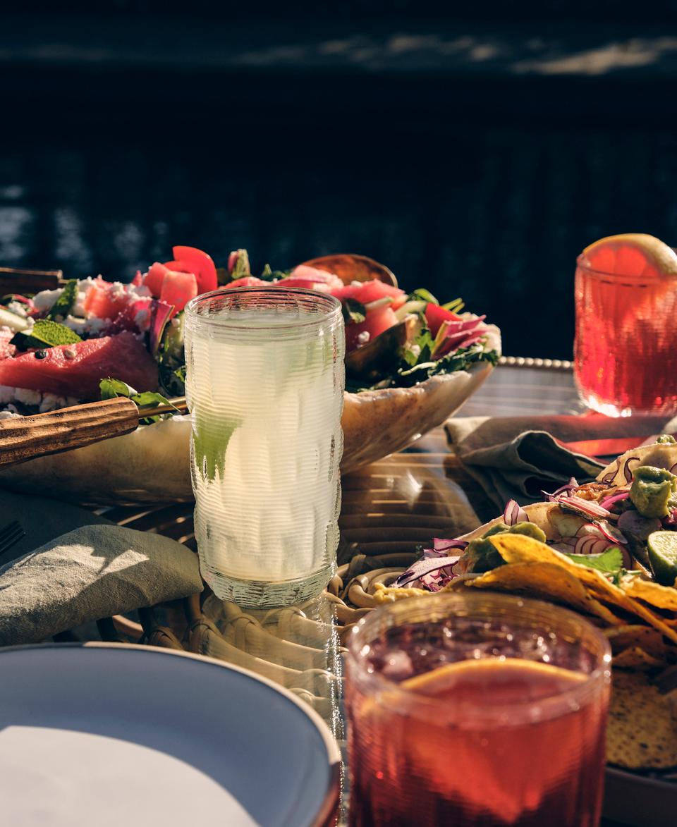 A table setup with plates of salad, tacos, and various drinks, including a glass of lemonade, in an outdoor dining setting with soft sunlight.