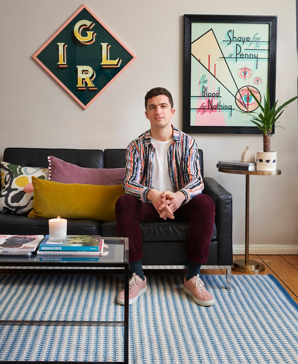 A person sits on a black sofa, surrounded by colorful pillows, a candle, and books on a glass coffee table in a stylish, art-decorated living room. Wall text: "IGLR," "Shave for a Penny let Blood for Nothing."