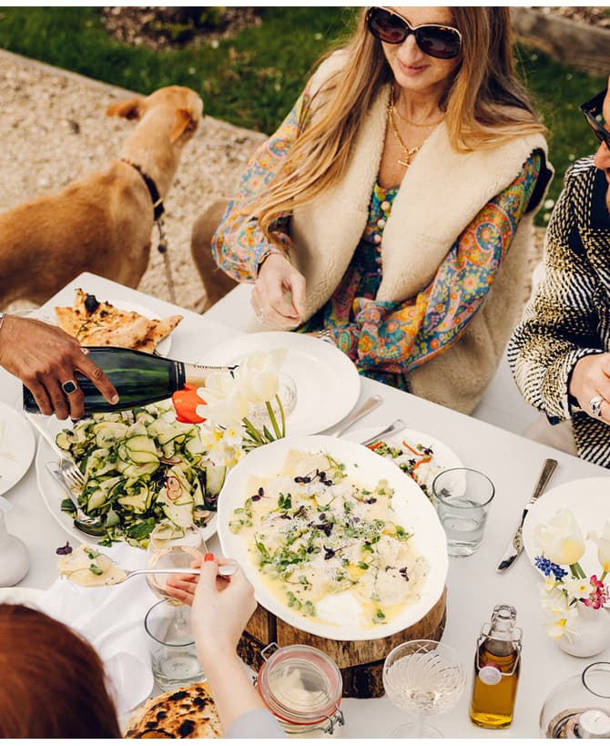 Several people dine outdoors, one pouring wine. The table is adorned with plates of food, flowers, and drinks. A dog stands beside them. The environment is grassy with gravel paths.