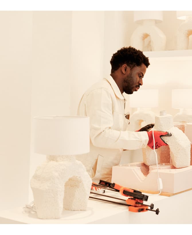 A person wearing gloves shapes a textured white lamp base on a wooden table surrounded by identical lamps and tools in a minimalist, brightly lit room.