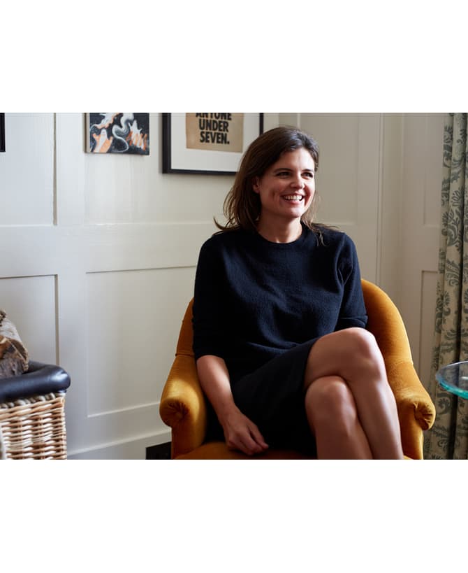 A woman sits smiling in a mustard-colored chair in a bright room with framed art, a wicker basket, wood logs, and patterned curtains. Text on artwork: "ANYONE UNDER SEVEN."