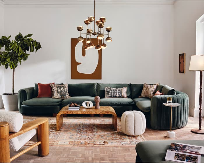Green sectional sofa with patterned cushions in a cozy living room, accompanied by wooden and modern furniture. A potted plant, floor lamp, and abstract wall art adorn the white walls.