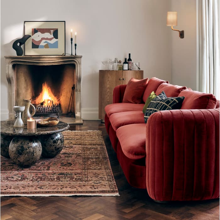 A red velvet sofa sits in front of a lit fireplace; a round marble coffee table holds decor items on an ornate rug in a cozy, well-lit living room with a chandelier overhead.