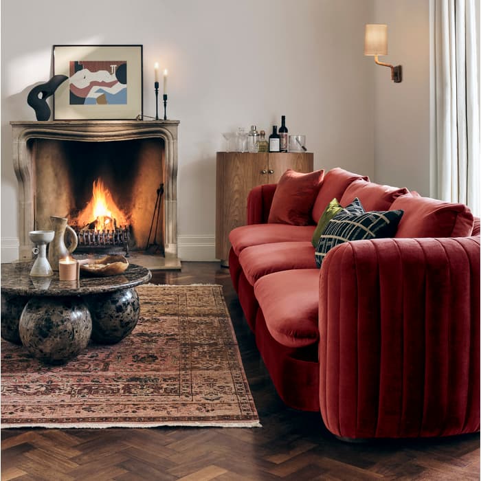 A red sofa sits in front of a lit fireplace surrounded by a rug, coffee table, artwork, and wine bottles, under a decorative chandelier in a cozy living room.