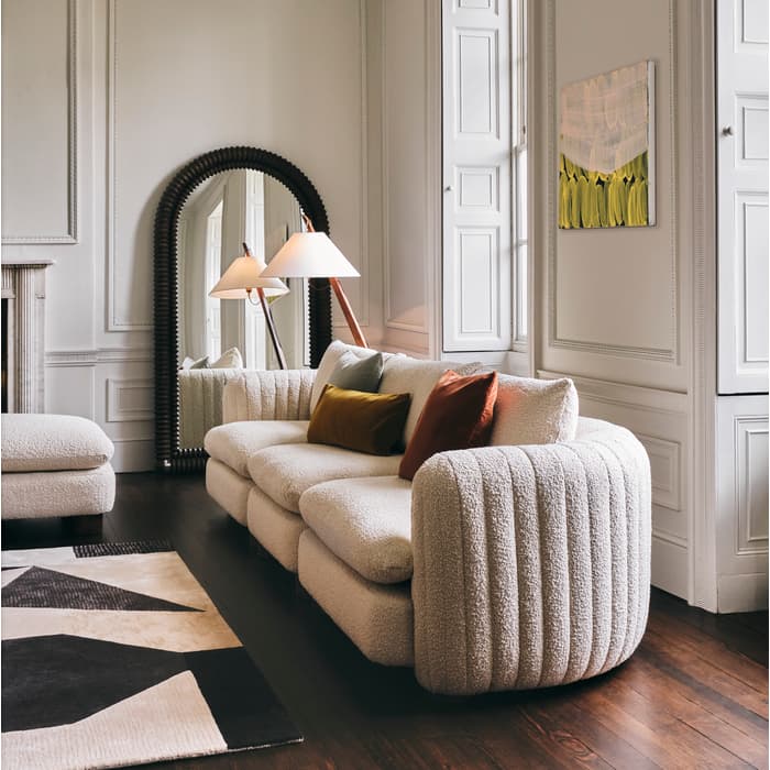 A cream-colored sofa with pillows sits in a stylish, minimalist room featuring white paneled walls, a large, ornate mirror, a geometric-patterned rug, a floor lamp, and a wall painting.