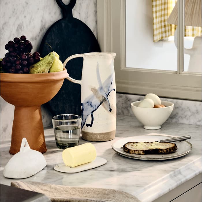Ceramic pitcher, fruit bowl, and butter dish are arranged on a marble countertop with a slice of buttered bread on a plate, against a background with a window and a lamp.