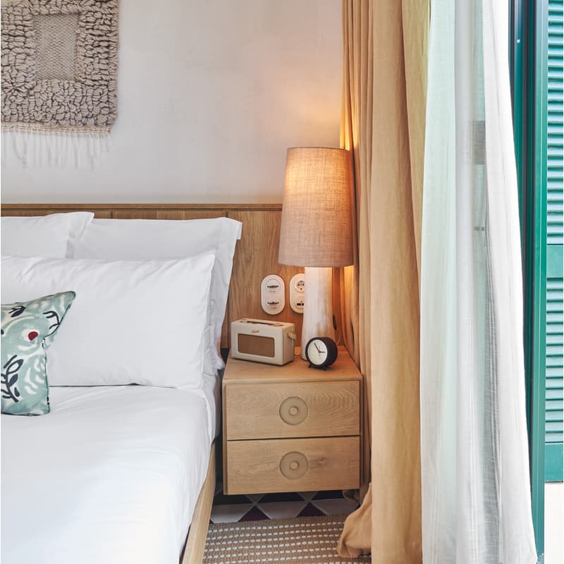 A double bed with white linens and a decorative pillow sits beside a wooden nightstand holding a lamp, clock, and radio, with curtains partly drawn revealing green shutters.
