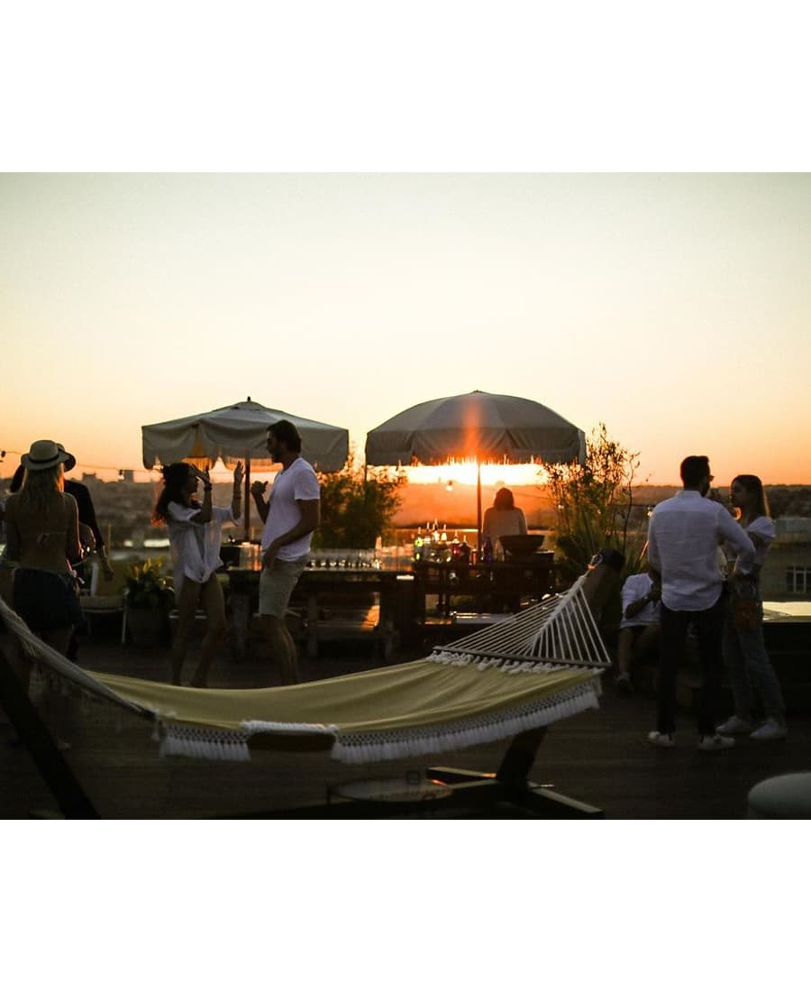 People are socializing and enjoying themselves on a rooftop at sunset, with a hammock in the foreground and umbrellas, lounge chairs, and a bar setup in the background.