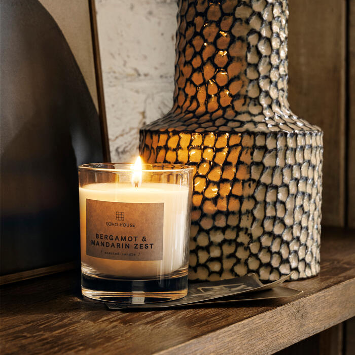 A lit candle labeled "SOHO HOUSE BERGAMOT & MANDARIN ZEST scented candle" sits on a wooden shelf beside a textured, patterned vase against a brick wall background.