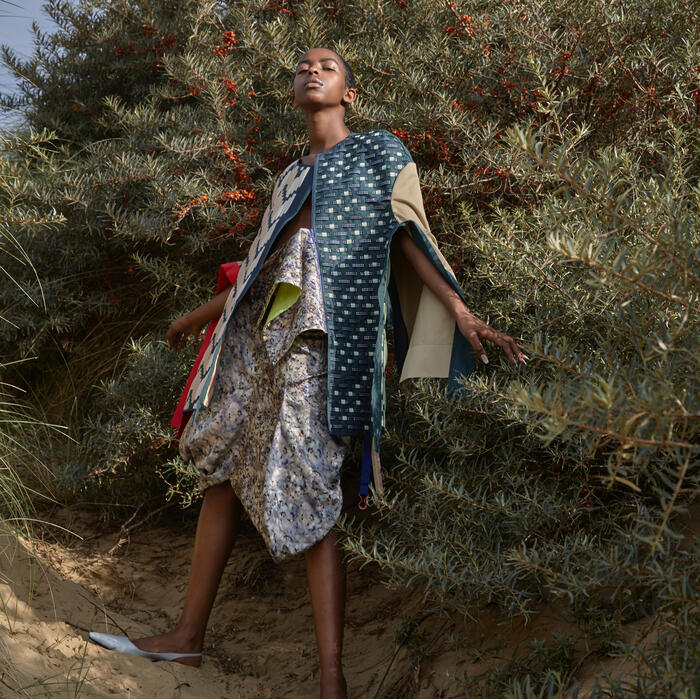 A person in colorful, textured clothing stands with arms outstretched among dense, green bushes with red berries on a sandy terrain, under a bright, cloudy sky.