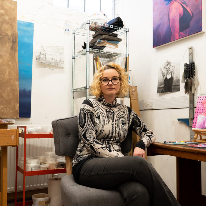 A woman with glasses sits on a chair in an art studio, surrounded by paintings, sketches, and art supplies on shelves and easels.