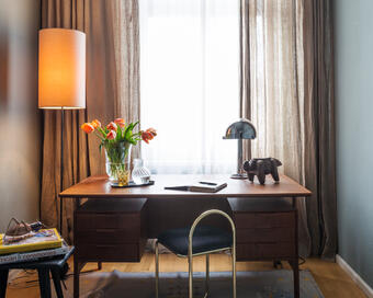 A wooden desk with flowers in a vase, a notebook, and a lamp is situated in a small room with a floor lamp, curtain-covered window, and a side table with books.