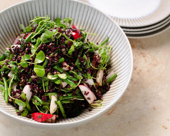 A textured white bowl filled with a vibrant salad consisting of mixed greens, black rice, green beans, and radishes sits on a neutral countertop beside a stack of white plates.