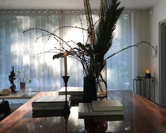 A vase with tall, decorative branches and leaves stands on a wooden table with a candle and books, including "Jan Schoonhoven," in a room with sheer curtains and ambient lighting.