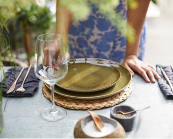 A person sets an outdoor dining table with green plates on a woven mat, next to folded napkins with cutlery and a wine glass, surrounded by greenery.