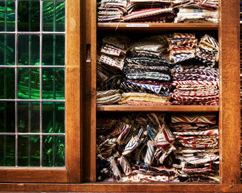 Stacks of colorful patterned fabric are neatly arranged on wooden shelves in an old cabinet with glass windows. A label reads "Parties - Bordados Muestrarios."