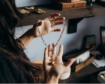 A person holds a gold chain necklace in a room with wooden shelves holding decorative items, such as a woven basket, antique lamp, and small trays.