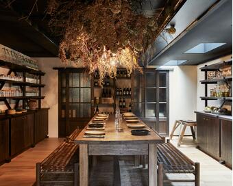 A long wooden dining table is set with plates and cutlery, surrounded by wicker chairs, in a warm, rustic dining room with shelves full of glassware and hanging dried plants.