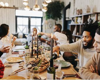 A group of people are enjoying a meal together around a wooden dining table, pouring drinks, and conversing in a warmly lit, cozy room with rustic decor.