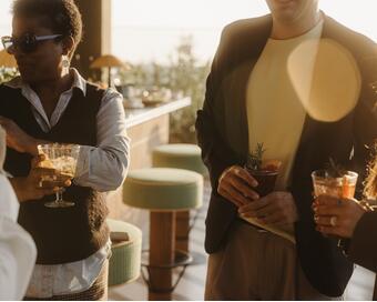 Three people holding cocktails, conversing and smiling in an outdoor setting during sunset, with green bar stools and plants visible in the background.