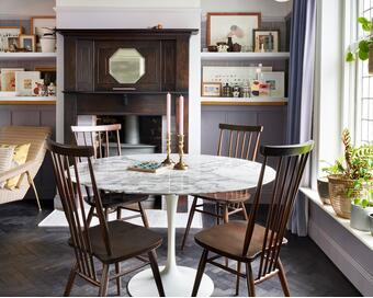 A round, marble-top table with wooden chairs and two tall candles is in a cozy room with a dark wood fireplace, framed art on shelves, and a large window with potted plants.