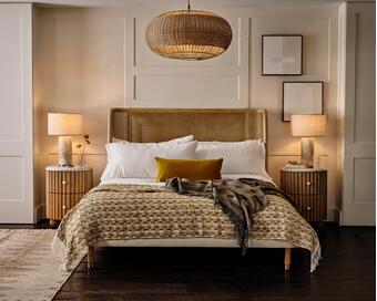 Bed positioned in the center with a mustard throw pillow and patterned blanket, flanked by two nightstands with lamps, in a stylish bedroom with wooden floors and minimalistic decor.