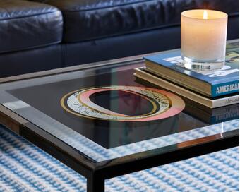 Candle and stacked books rest on a glass-top coffee table featuring decorative artwork, situated in front of a dark leather couch over a blue patterned rug. Text on books includes "AMERICAN YOUTH" and "Alexander McQueen."