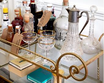 A gold bar cart holding assorted bottles with labels, glassware, and books, is set against a white paneled wall in a room with a brown floor. Labels read: "Aperol 28.8.18", "30.05.18," and "HONEY SMOKE SCOTCH."