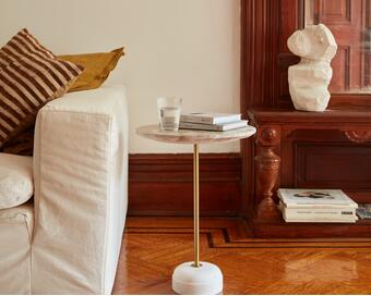 A round side table with a glass of water, stacked books, and a beige couch with striped and solid cushions in a cozy living room near an ornate wooden cabinet holding a sculpture.