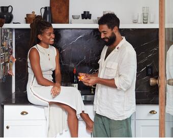 A woman sits on a kitchen counter smiling at a man in loose, light clothing who peels an orange. They are in a modern kitchen with dark countertops and a shelf with dishes.