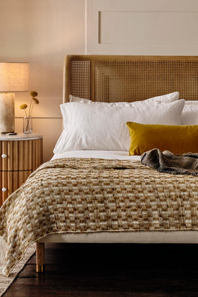 A neatly-made bed with a yellow pillow and checkered blanket stands between two wooden nightstands with lamps, set against a cream-colored paneled wall with two abstract artworks, in a modern bedroom.