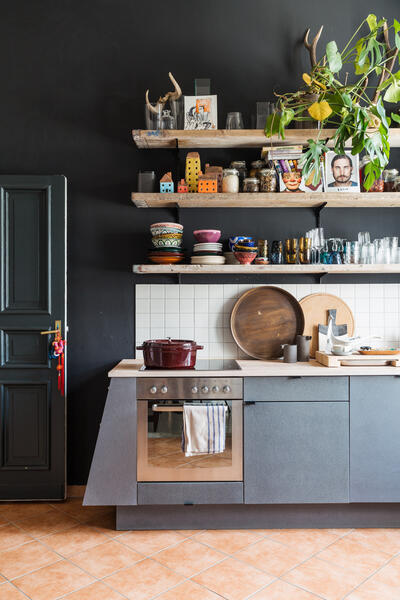 A kitchen with a gray oven below a counter holding a red pot, surrounded by plates and cutting boards; wooden shelves above display decorative items, jars, and glasses against a dark wall.