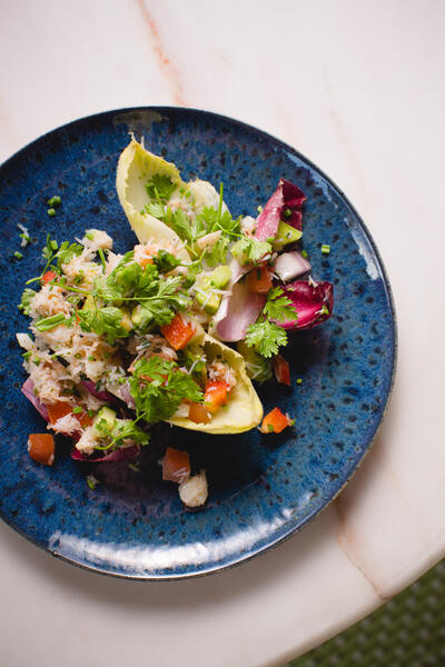 A blue plate holds a colorful crab salad with mixed greens, diced tomatoes, onions, and herbs atop a marble table.