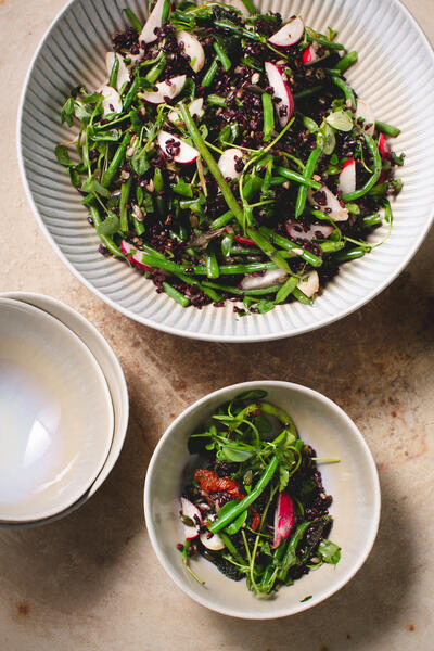 A large bowl of black rice and green bean salad with radishes and greens next to stacked empty bowls on a light-colored wooden surface.