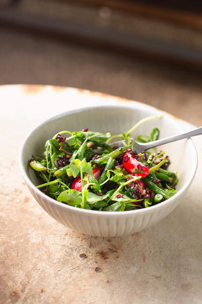 A white bowl contains a fresh green salad with slices of tomatoes and dark grains, sitting on a textured, rustic table. A fork is placed in the bowl, ready for use.