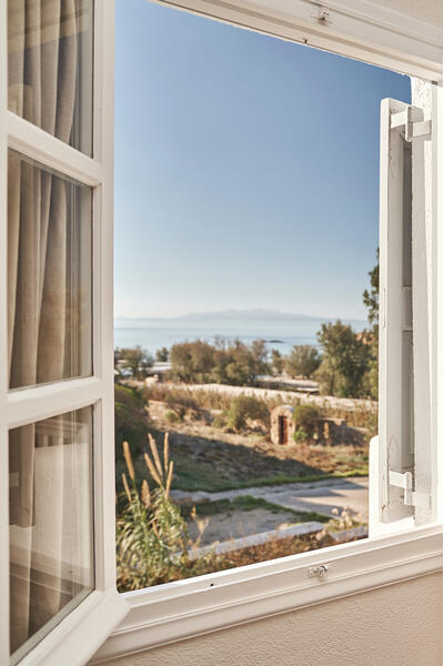 Open white window overlooks a picturesque landscape with trees and distant mountains by the sea; inside, books and a candle holder sit on a small cabinet.