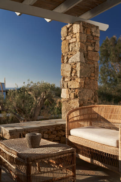 Wicker furniture (a cushioned seat and table) sits under a wooden pergola beside a stone column, overlooking a lush garden with a glimpse of the ocean in the background.