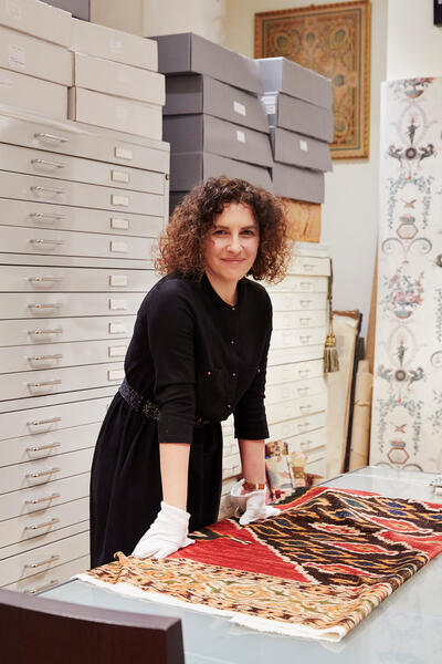 A woman wearing white gloves examines a richly patterned fabric on a table, surrounded by storage drawers and boxes in an organized archival room with decorative wall art.