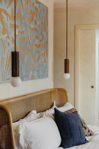 A rattan headboard frames pillows on a bed. Two pendant lights hang above near an abstract, blue and orange wall art piece in a softly lit bedroom.