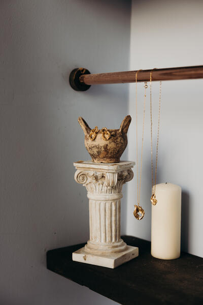 Gold jewelry hanging from a rusty pipe and an ancient-style vase atop a white column; next to it stands a white candle on a dark surface against a wall.