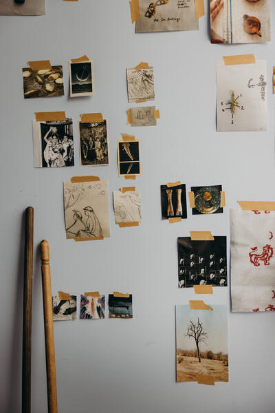 Photos and sketches are taped to a white wall in a casual arrangement. Below them, wooden poles lean against the wall. Different textures and colors create a collage-like display.