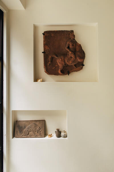 Ancient terracotta relief depicting a horse rider and a carved stone plaque with a panther, displayed on recessed shelves in a minimalist, cream-colored wall. Small vases and figurines accompany.