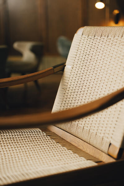 A wicker chair with wooden armrests is shown in close-up, situated in a warmly lit, stylish room furnished with other contemporary chairs in the background.