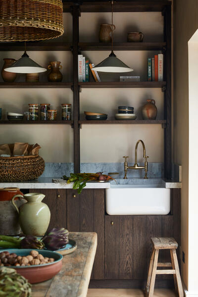 Kitchen sink occupies a marble countertop with a brass faucet, under dark wooden shelves holding books and pottery, surrounded by baskets, jars, and bowls; fresh vegetables lie nearby.