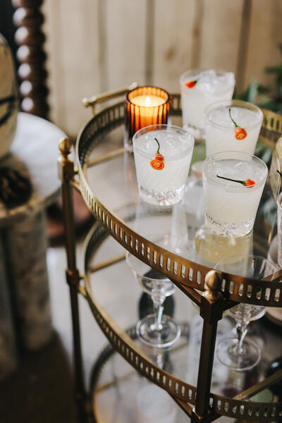 Four glasses of iced cocktails with red chilies sit on a gold, two-tier serving cart near a small candle; they are in an indoor setting with a marble side table nearby.