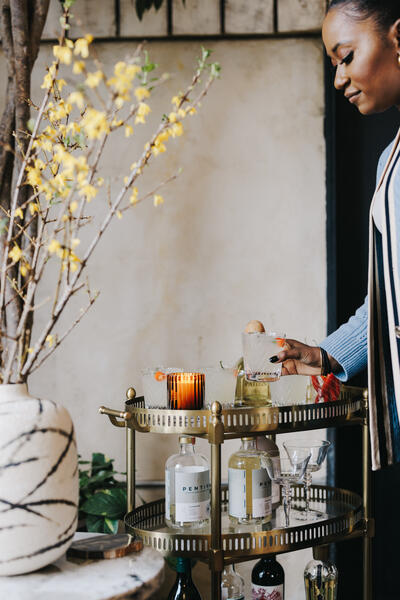 A person picks up a drink from a brass bar cart holding various beverage bottles and glasses, situated beside a potted plant with yellow flowers in a warmly lit indoor setting.